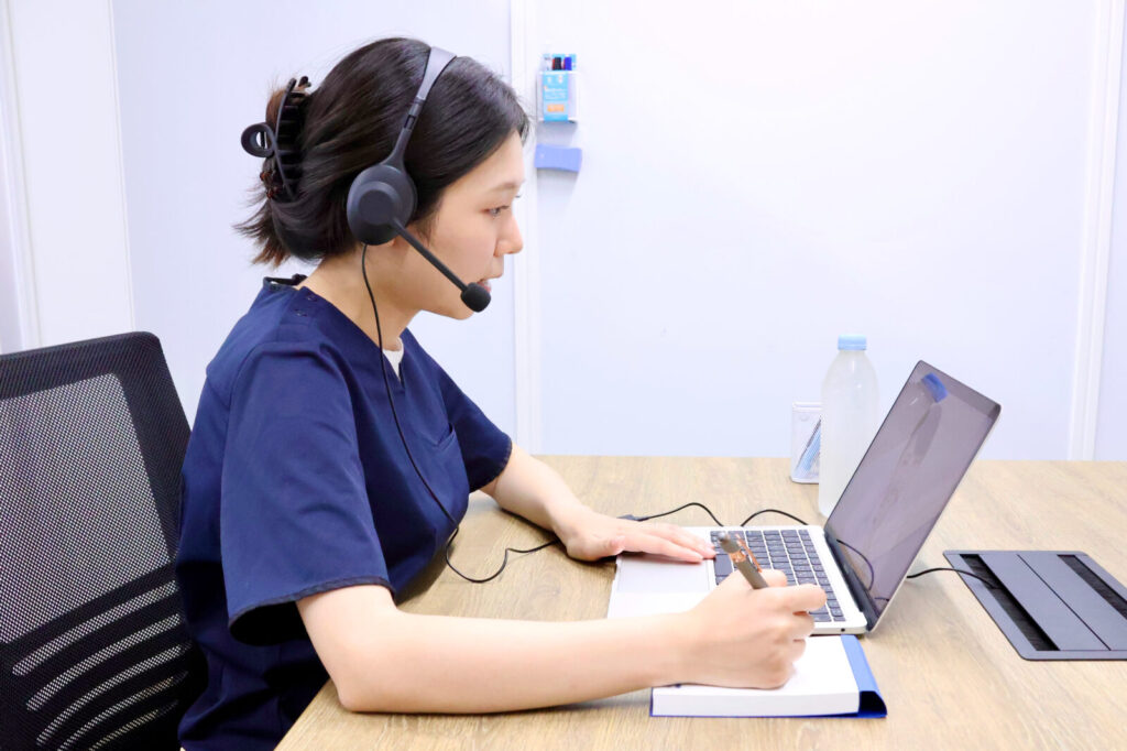 ドクターメイトのオンコールナースが、室内でPCの前に座りオンラインでやり取りをしている様子を横から写した写真。オンコールんナースはネイビーのナース服を着用し、耳元にはインカムをつけています