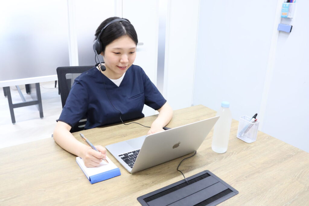 デスクでノートパソコンを開き、画面に向かって笑顔で話しかけているドクターメイトのオンコールナースの女性の写真。インカムをつけ、左手でパソコンを操作しながら、右手ではペンを持ってメモを取っています。